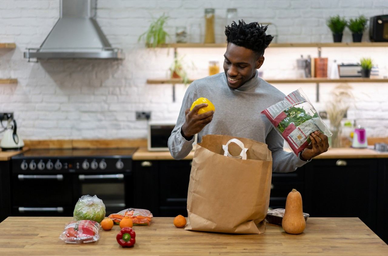 Man holding a fruit in his hand while standing at the kitchen with grocery bag opened